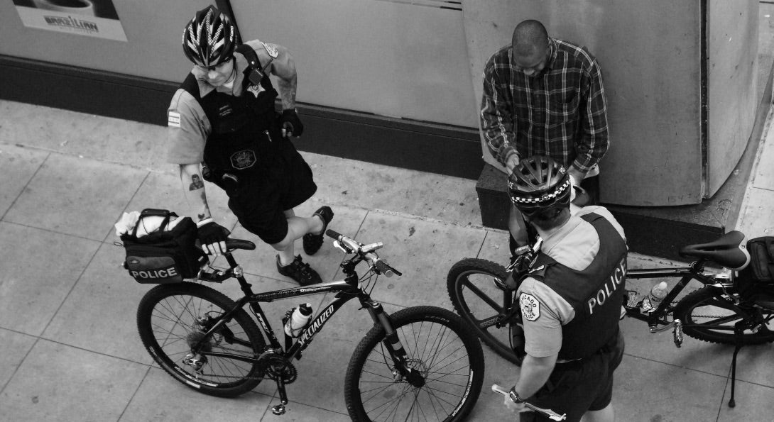 Two Chicago police officers, who have been on bikes, talk with a Black man standing on a street corner in downtown Chicago.